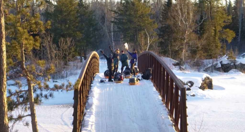 a group of cross country skiers, each pulling small sleds, make their way across a snowy bridge